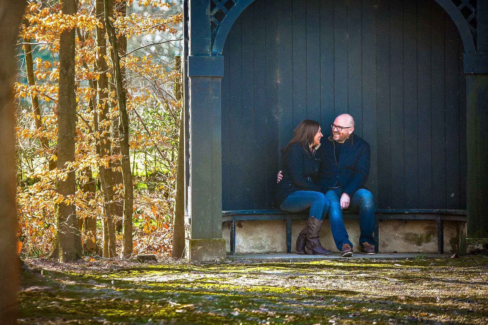 Couple sitting together in a wooden shelter surrounded by autumn woodland, engagement portrait photography by Hector's Eye