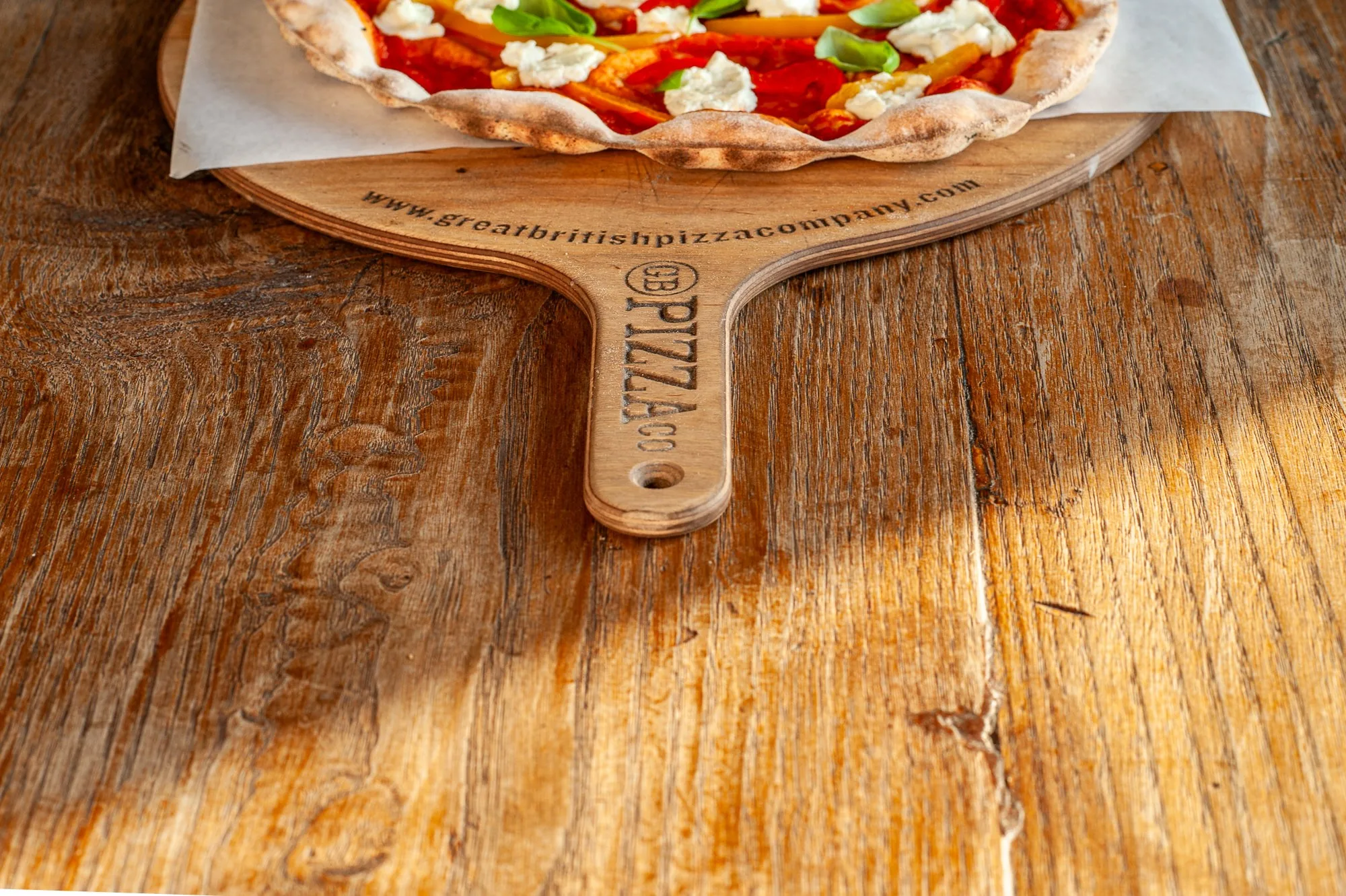 Wood-fired pizza with peppers and goat's cheese resting on a branded pizza peel, food brand photography by Hector's Eye